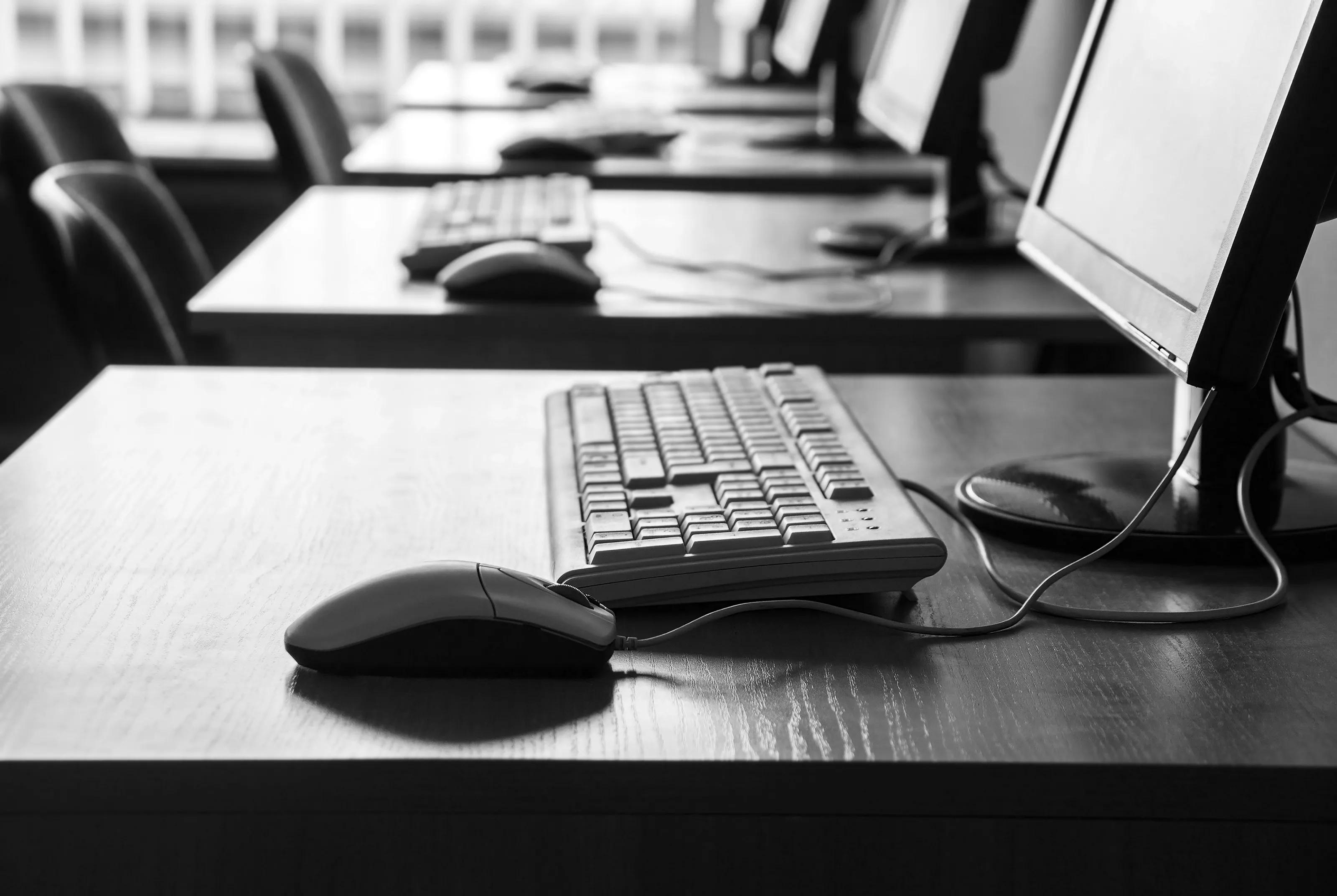 Row of workplaces - four wooden tables with computers (monitors, keyboards and mouses) by a window in an office or classroom in black and white.