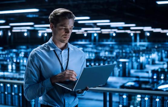 Handsome Smiling IT Specialist Using Laptop Computer in Data Center. Succesful Businessman and e-Business Entrepreneur Overlooking Server Farm Cloud Computing Facility.