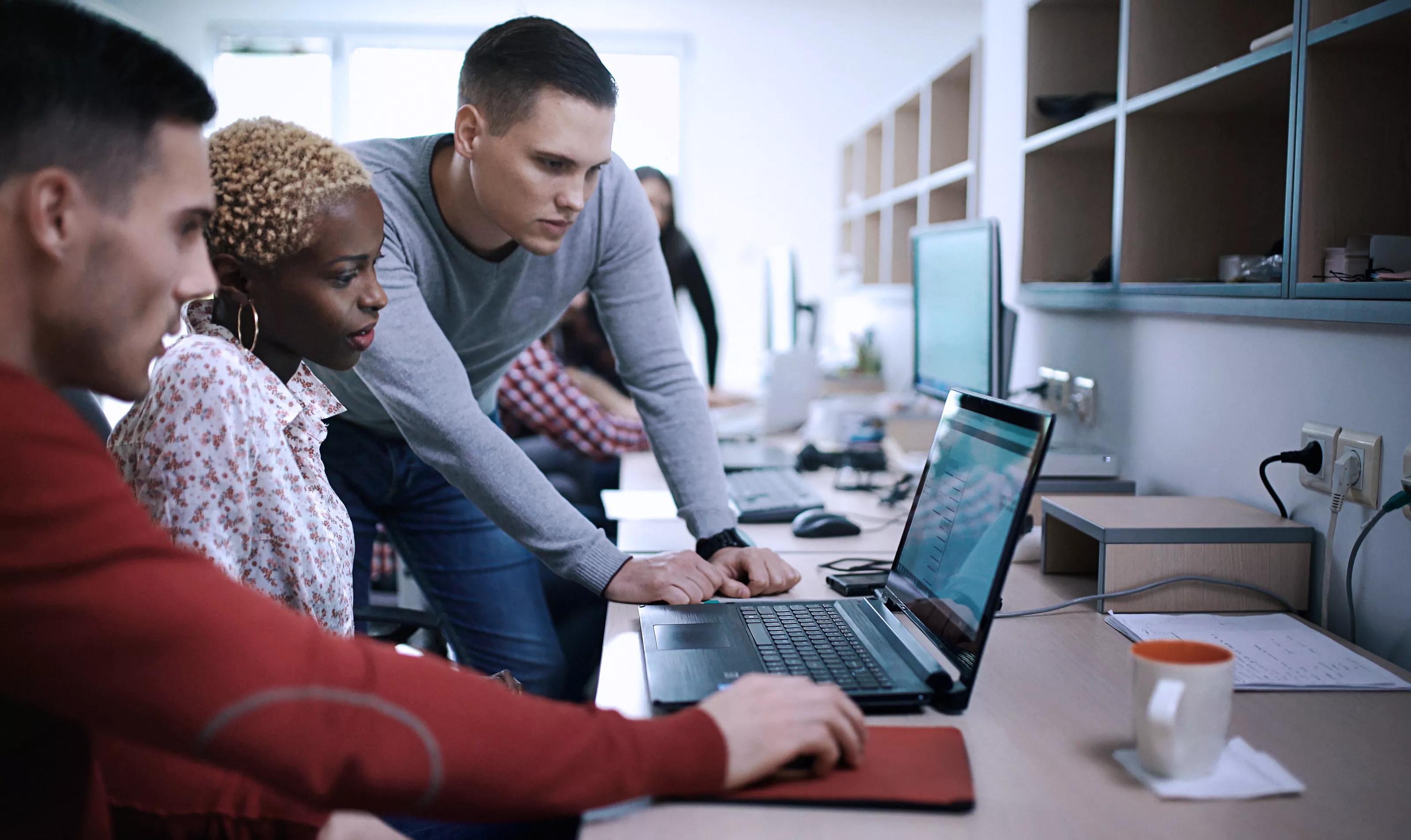 Closeup of group of application developers discussing about certain code for an application. There's black woman two caucasian men gathered around a desktop computer. They are late 20's, mixed race, dressed smart casually. Blurry people in background, also released.