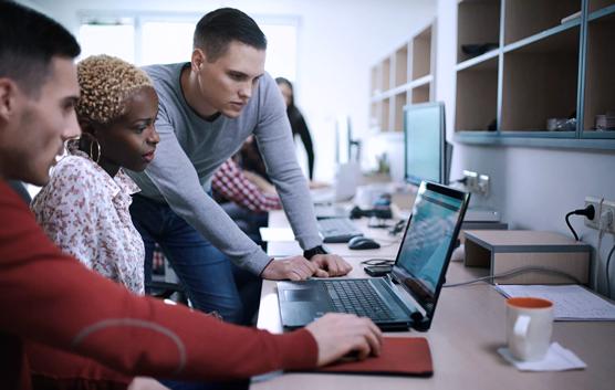 Closeup of group of application developers discussing about certain code for an application. There's black woman two caucasian men gathered around a desktop computer. They are late 20's, mixed race, dressed smart casually. Blurry people in background, also released.