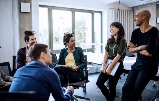 Colleagues looking at cheerful businesswoman holding laptop in meeting at creative office