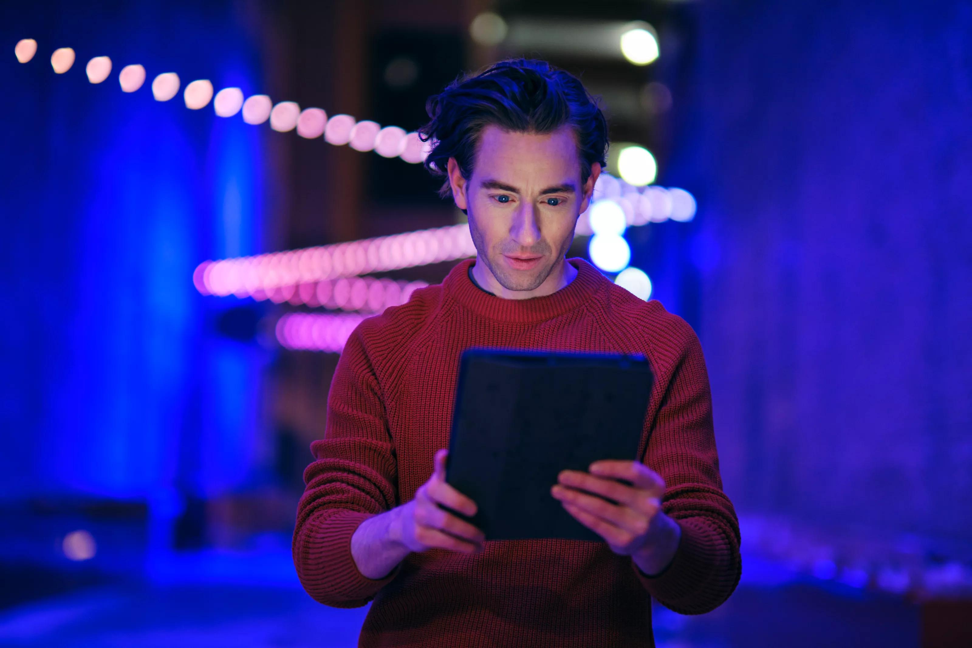 A young man using a digital tablet at night in a city with blurred lights.