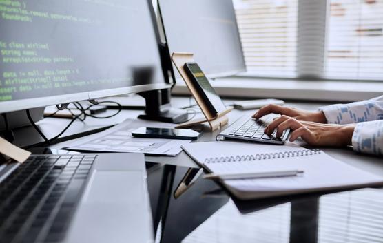 Close-up of female programmer typing code on keyboard from smartphone while sitting at table in front of computer monitors at office