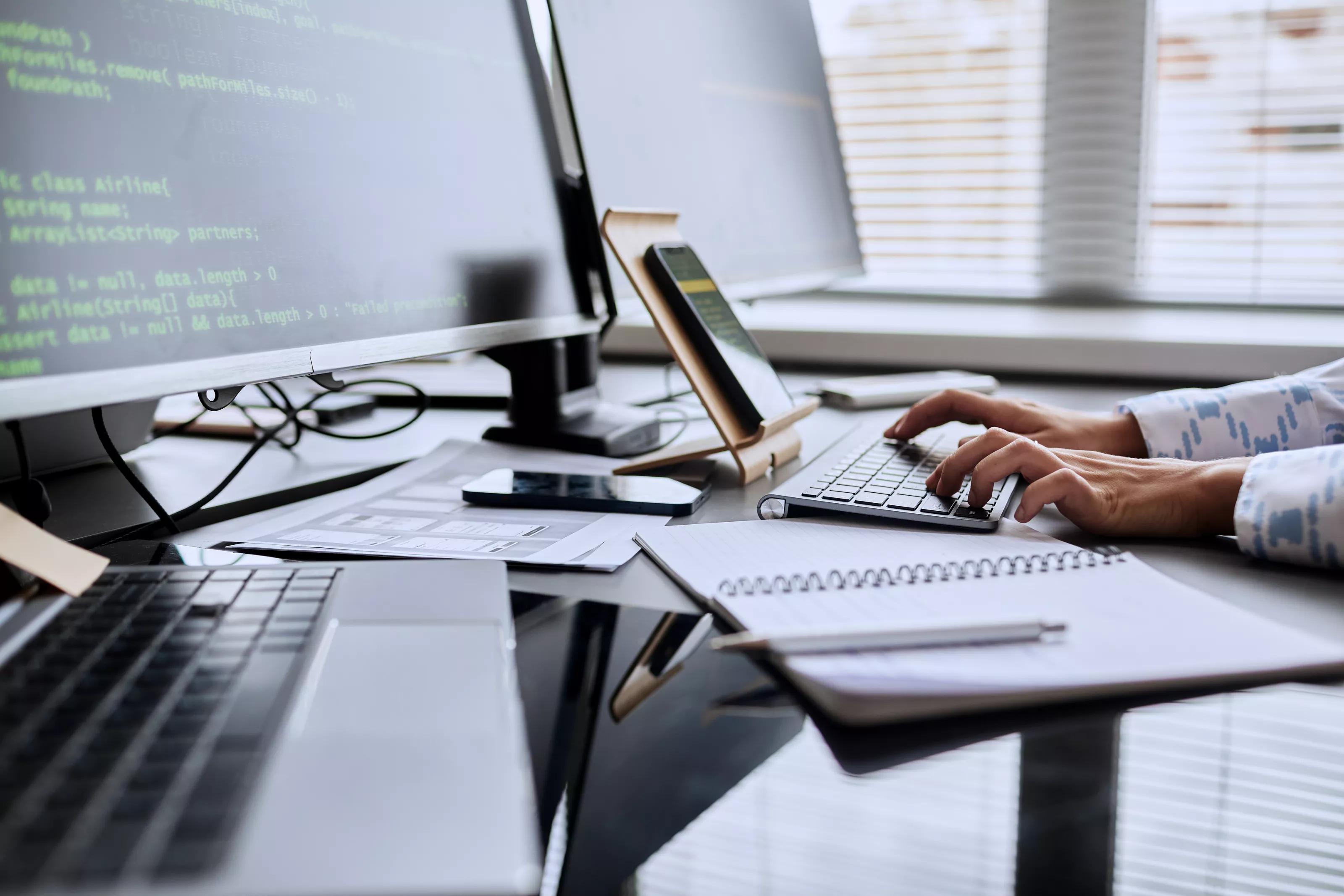 Close-up of female programmer typing code on keyboard from smartphone while sitting at table in front of computer monitors at office
