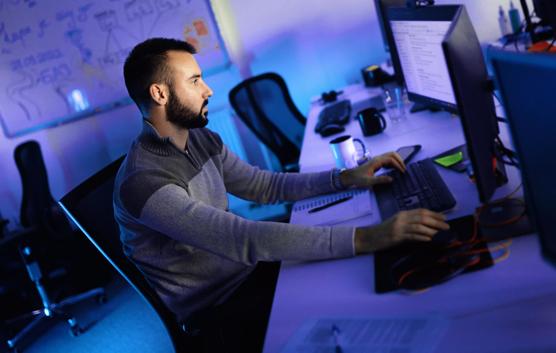 Closeup side view of a young man coding on multiple computers early in the morning.