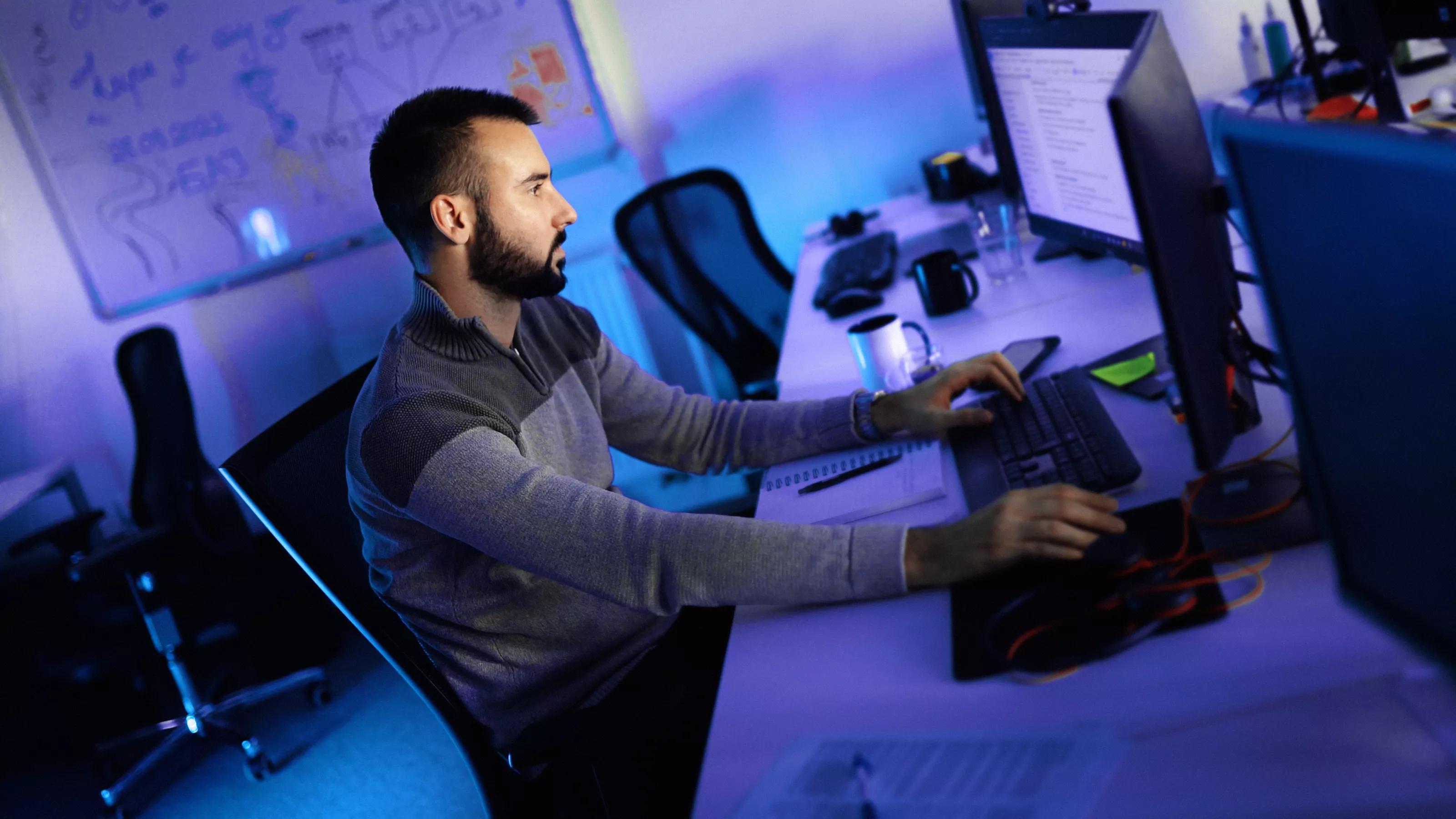 Closeup side view of a young man coding on multiple computers early in the morning.